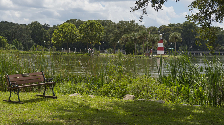 View of the red and white striped lighthouse on marshy Lake Dora from a public park. A bench is situated for people to sit on in the foreground.