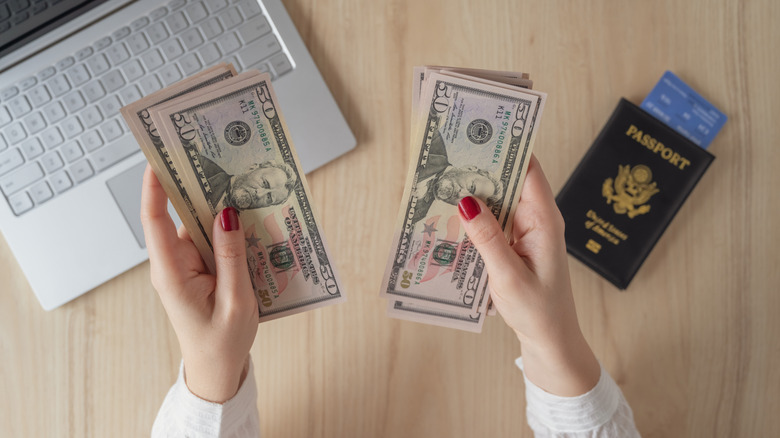 Flat lay of woman's hands holding stacks of $50 bills with an American passport, boarding pass, and laptop on the table