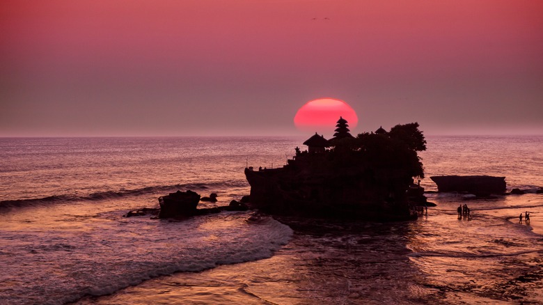 Tanah Lot temple, Bali, Indonesia, tourists walking in the ocean at sunset, huge red sun in background and backlit silhouette