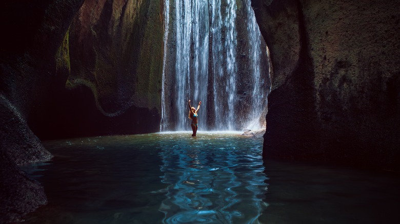 Woman stands in underground cave pool under falling fresh water of Tukad Cepung waterfall. Nature day tour, hiking activity adventure and fun at family tourist camp on summer vacation in Bali island