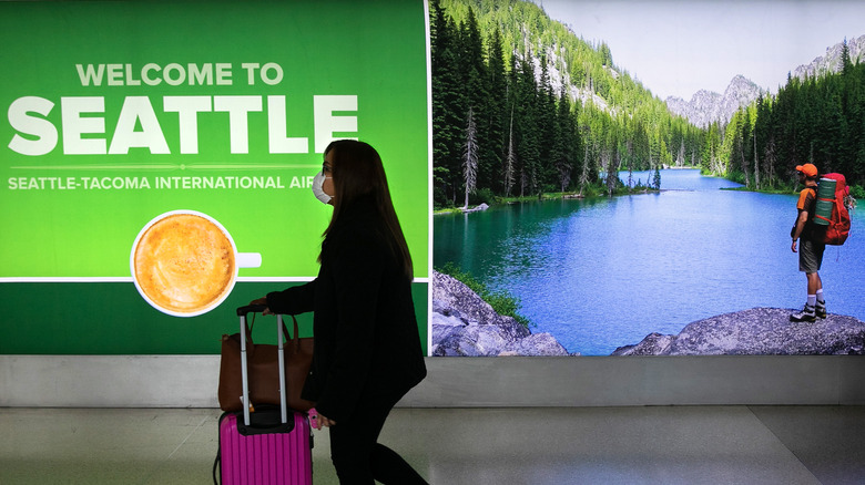 Woman wearing mask walks past "Welcome to Seattle" sign at the airport.