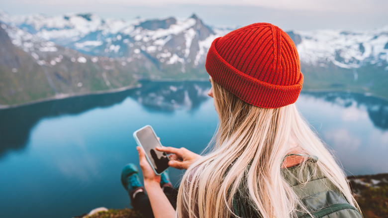 hiker on their phone with snow capped mountains and a lake in the background