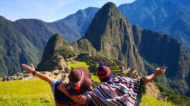 Two people in ponchos throwing their arms out in front of an aerial view of Machu Picchu, Peru