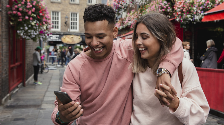 Couple with their arms around each other standing outside flowered pub smiling while looking at a phone
