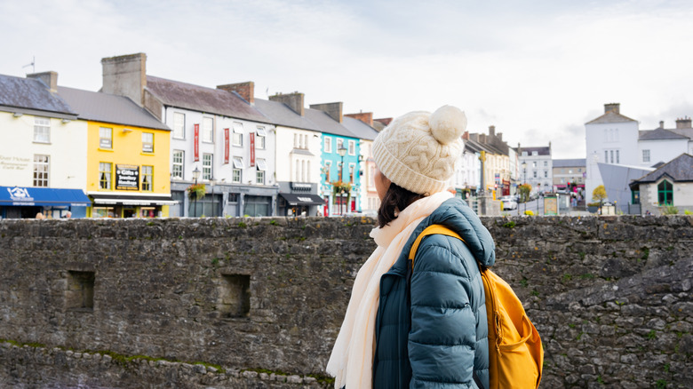 Dark haired woman in a hat, coat, scarf and yellow backpack in front of colorful row houses