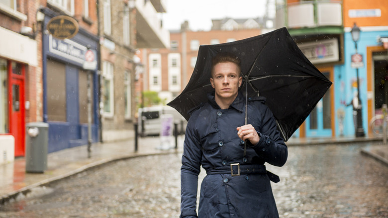 Redheaded man walking down an Irish street with an umbrella wearing a blue trenchcoat