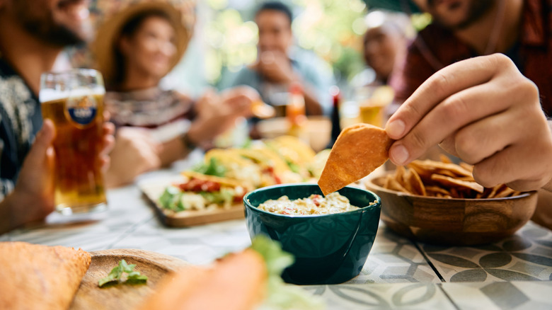 A tortilla chip is dipped at a Mexican restaurant table with people dining in the background.