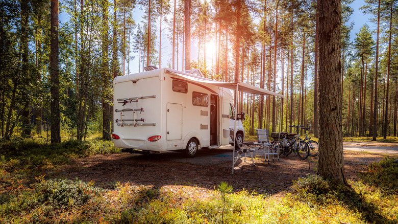 A white RV is pictured in a forest with an awning, camping chairs, and bicycles.