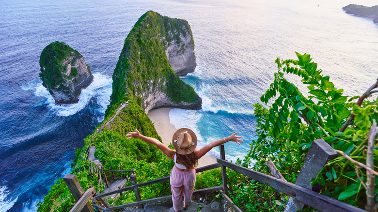 Woman in straw hat stands with her arms out on steep steps near dramatic rock formations on the coast of Nusa Penida, Indonesia