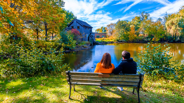 Man and woman sit on a wooden bench overlooking a pond and historic mill in Illinois during early autumn