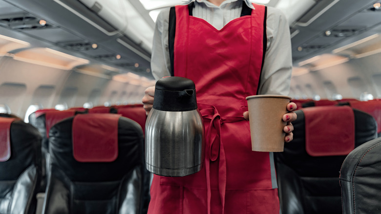 A flight attendant stands in the airplane aisle holding a hot liquid carafe and a paper cup.