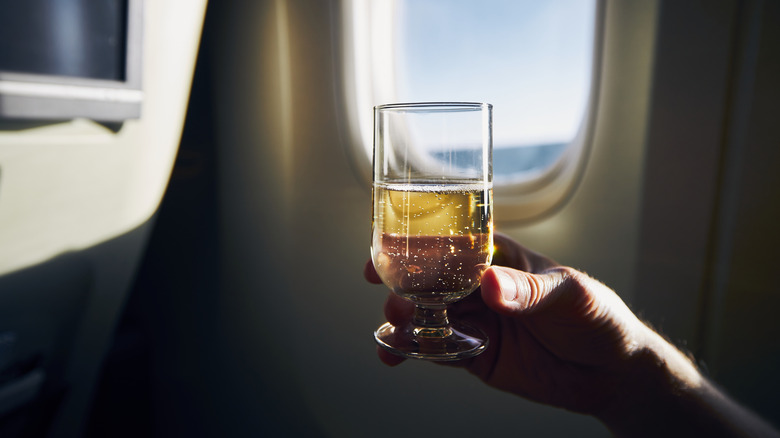 A passenger raises a glass of sparkling wine in front of an airplane window mid-flight.