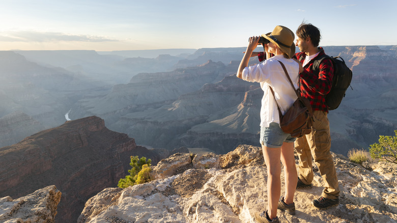 A couple of hikers stand high on a ledge overlooking the Grand Canyon with binoculars