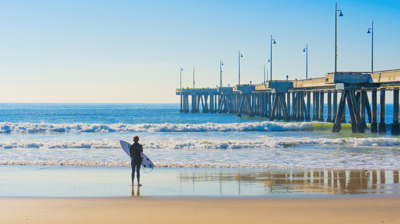 person holding a surf board standing at water's edge next to a pier