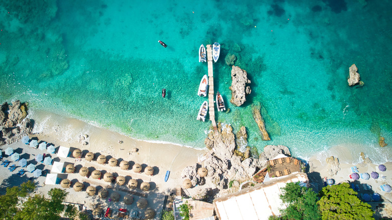 Aerial view of Dhermi Beach near Himarë on the Albanian Riviera, with blue water, white sands, umbrellas, and boats at a pier