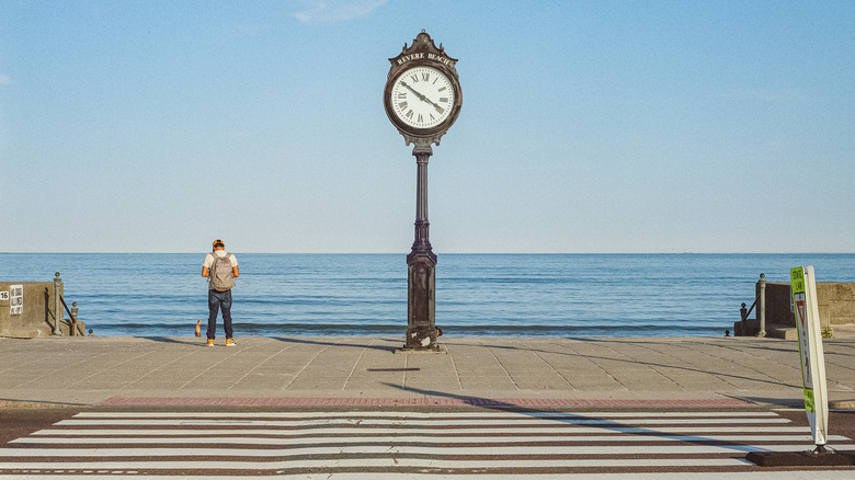 A man stands on the boardwalk near Revere Beach near a clock with the ocean in the background.
