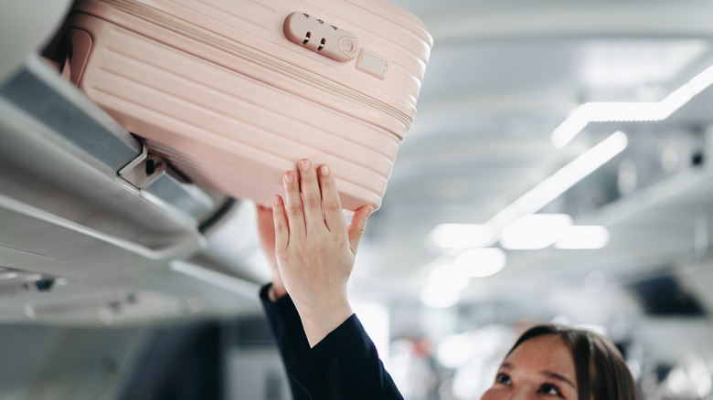 A woman placing pink carry on bag into overhead storage compartment inside airplane cabin preparing for travel.