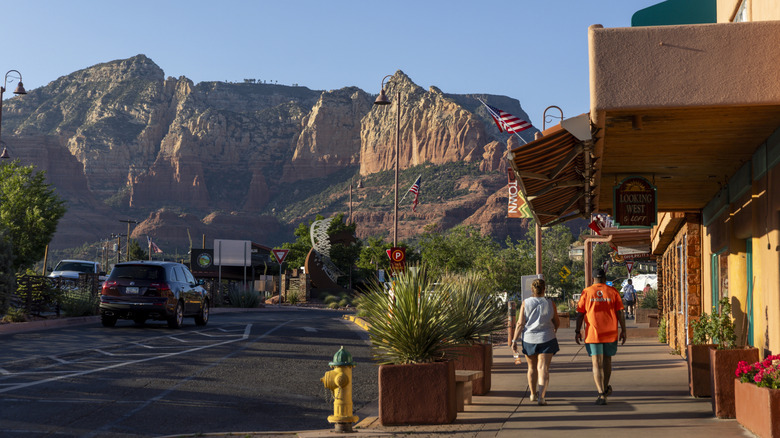 Two people walking on a sidewalk in front of a block of shops with sandstone mountains in the background