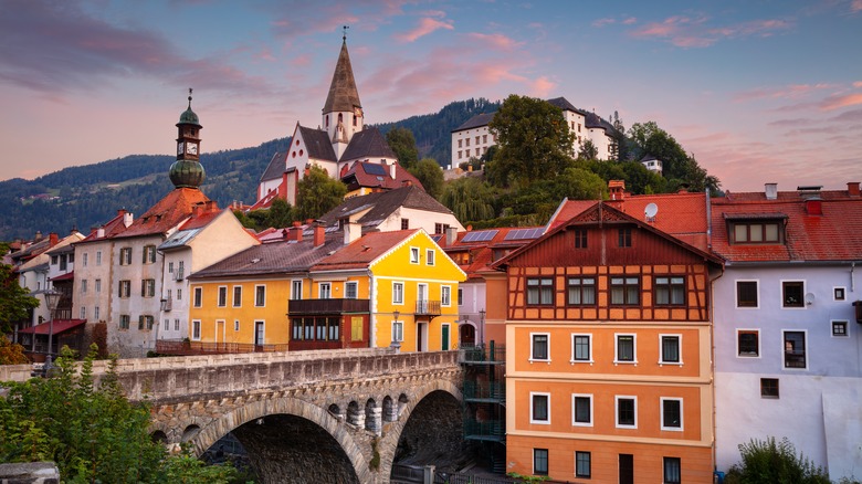 View of traditional buildings and a stone bridge in an Austrian village at sunset, with the Alps in the background