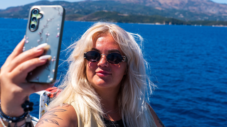 A woman on a boat, taking a selfie with a smartphone, with the blue water behind her