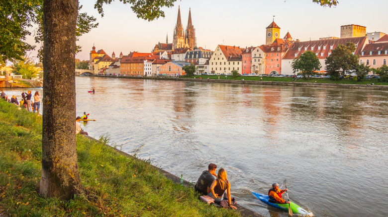 People sitting on grassy riverbanks with a view of Regensburg, Germany's Old Town