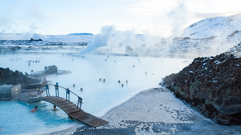 Wooden bridge over the milky blue thermal waters of Iceland's Blue Lagoon with steam, swimmers, and black lava landscape
