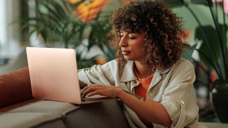 A woman sits at a laptop with plants in the background.