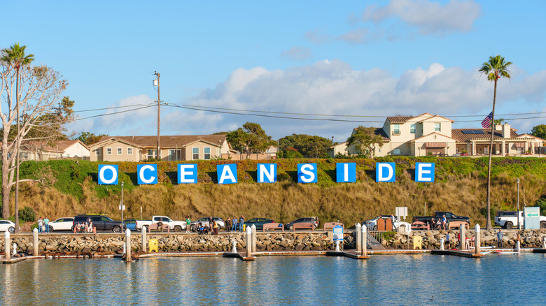 Letters spelling "Oceanside" on a grassy cliff near the water, with cars parked in front