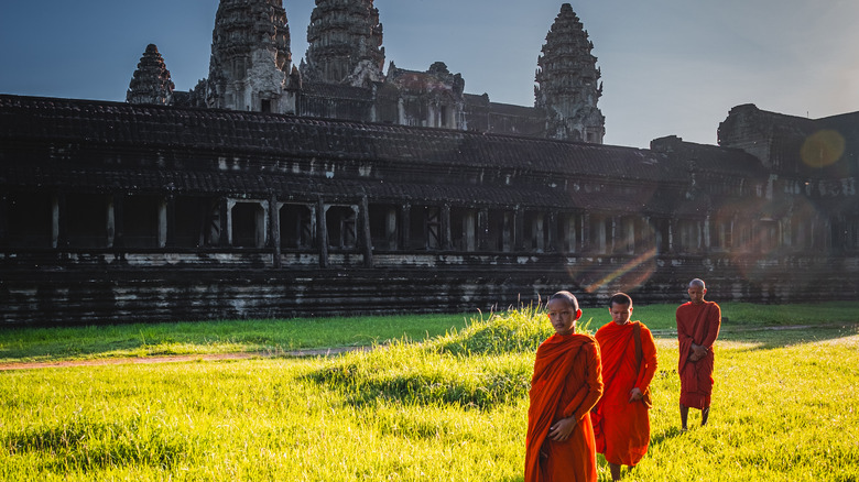 Three orange-robed monks walking away from the beehive towers of Angkor Wat on a grassy field, Cambodia