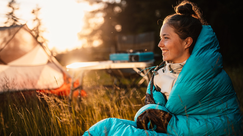 A smiling woman wrapped in a sleeping bag sits in the grass at a campsite.