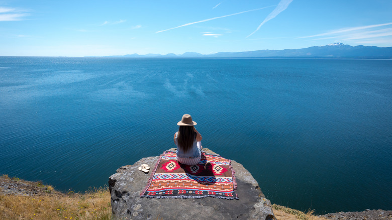 Woman on a red patterned blanket perches on the edge of a rock overlooking the Pacific from Hornby Island, BC, Canada, on a sunny day