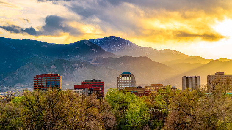 Buildings rise above the tree line in Colorado Springs at sunrise with mountains in the background.