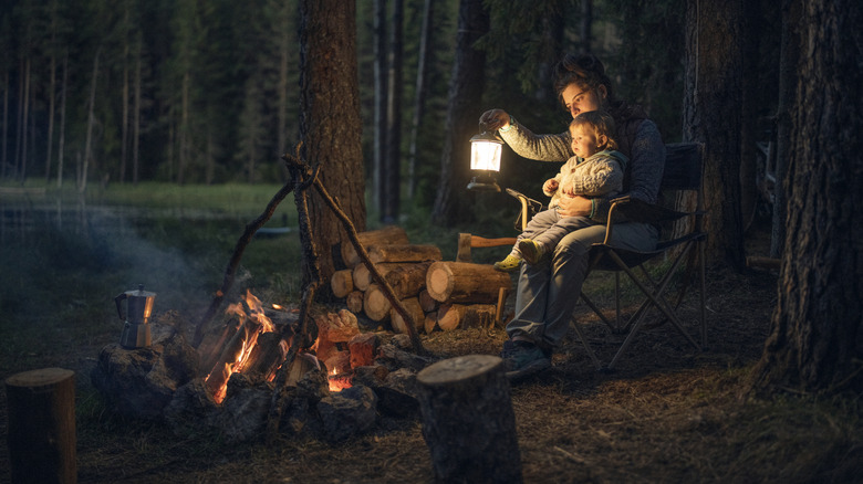 A mother holds a lantern and her young baby, both seated in front of a campfire at night.
