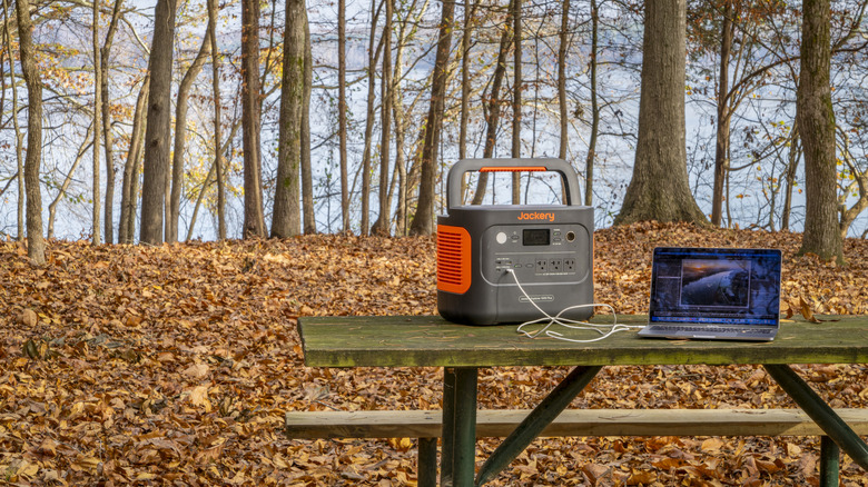The Jackery Explorer 1000 Plus charges a MacBook Pro laptop on a picnic table with dry autumn leaves on the ground.