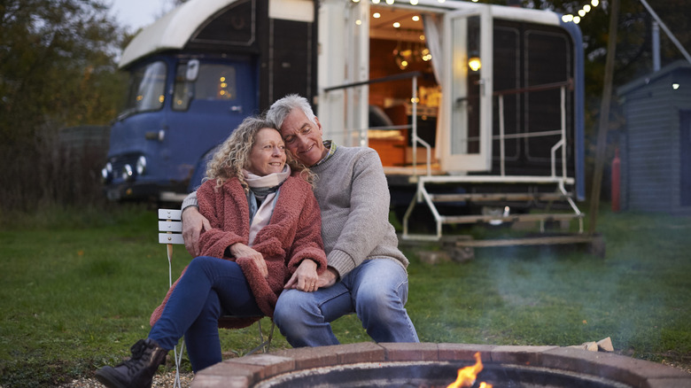 An older couple dressed in layers embraces in front of a campfire, with a blue retro-style camper situated in the background.