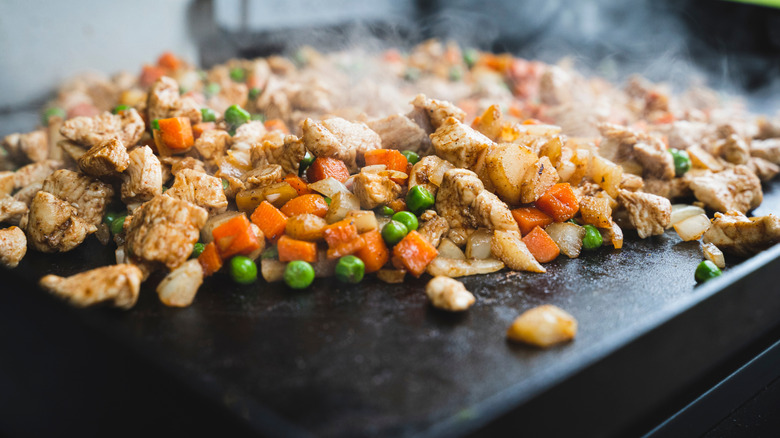Peas, carrots, potatoes, onions, and meat cook on an outdoor camping griddle.