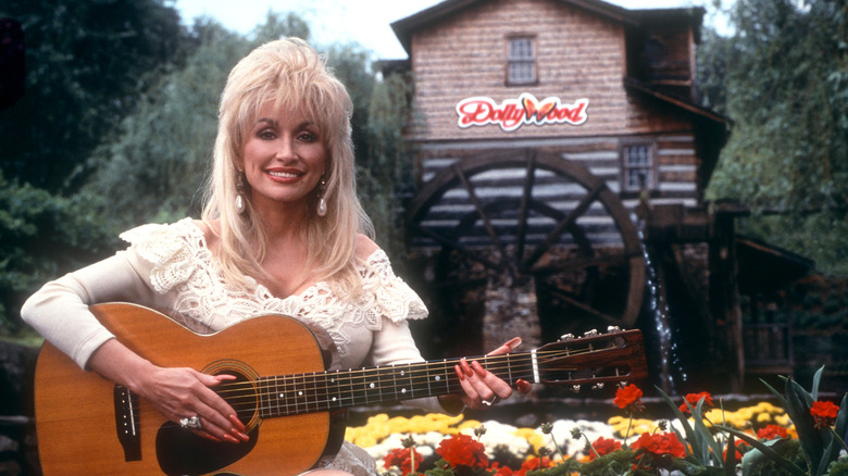 Dolly Parton at Dollywood, smiling and holding an acoustic guitar in front of a building