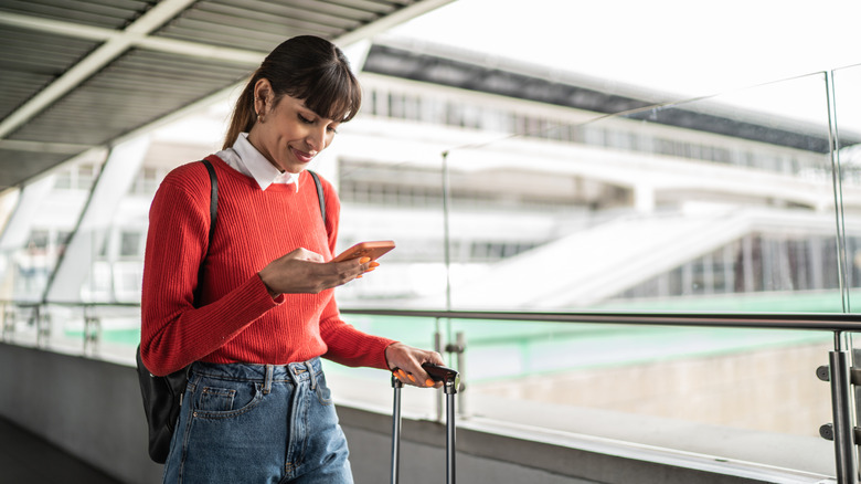 A woman checks her smartphone while walking her luggage through an airport.