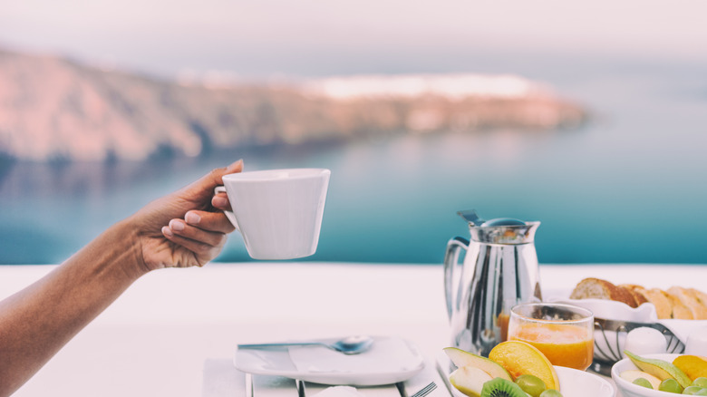 A hand holding a coffee cup over a luxurious breakfast table of fruit and bread overlooking the water of Santorini, Greece