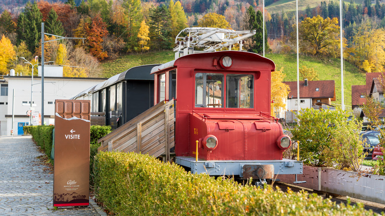 Old-fashioned red train engine near a visitor's sign, surrounded by autumnal foliage and outbuildings, at the Maison Cailler Chocolate Factory in Broc, Switzerland
