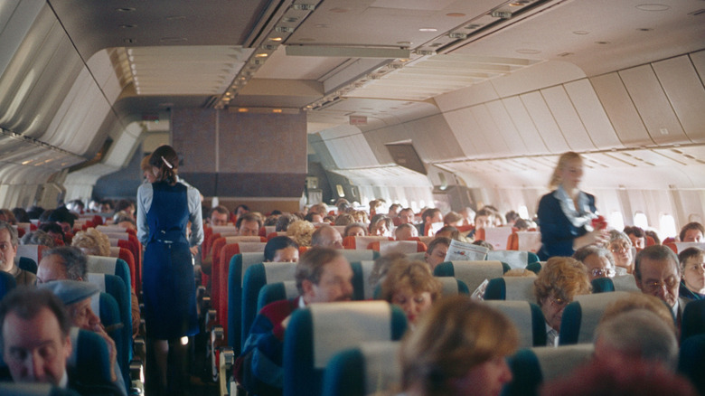 Two flight attendants in blue traverse the aisles of a retro plane full of people in the 1980s