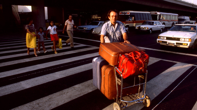 Man in a blue polo shirt crosses a crosswalk with a luggage cart full of brown suitcases in the 1980s.
