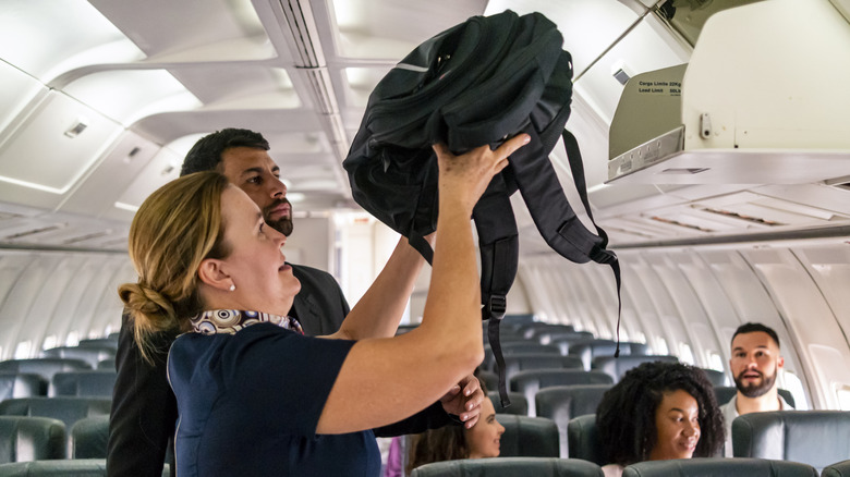 A flight attendant assists a passenger with stowing a backpack in an overhead bin during boarding.