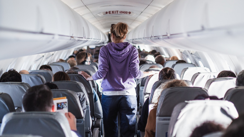 A female passenger is pictured from behind and walks the aisle of a fully seated airplane.