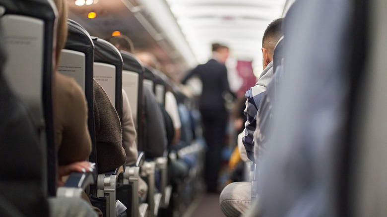Passengers sit on an airplane while a flight attendant stands in the aisle in the unfocused background.