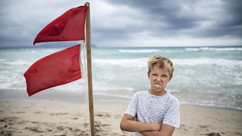 Angry young boy standing on the beach next to two red flags that mean no swimming allowed.