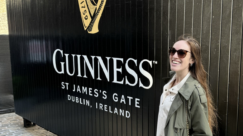Brunette woman in an olive trench coat posing by the famous black Guinness St James's Gate Dublin, Ireland gate