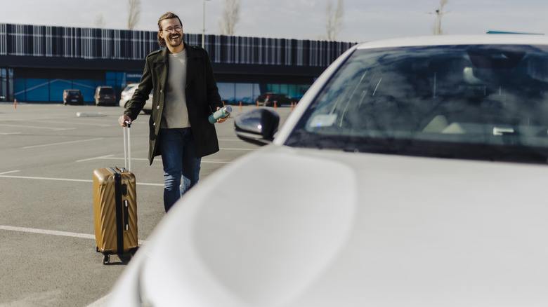 A smiling man walking to a car while rolling a suitcase
