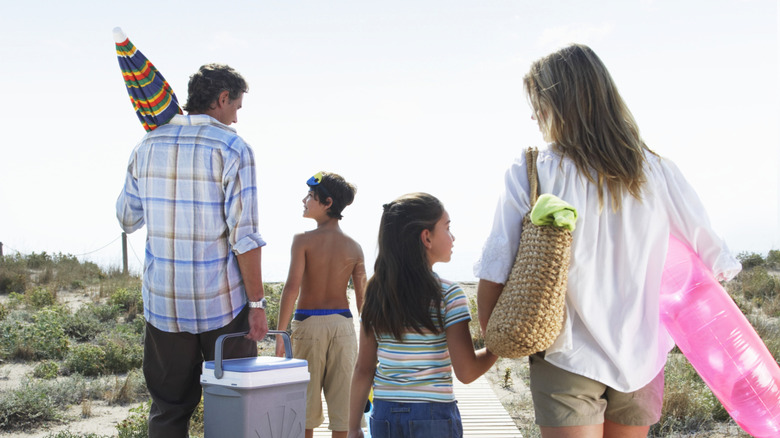 Parents with two kids approach the beach with cooler and float toys.