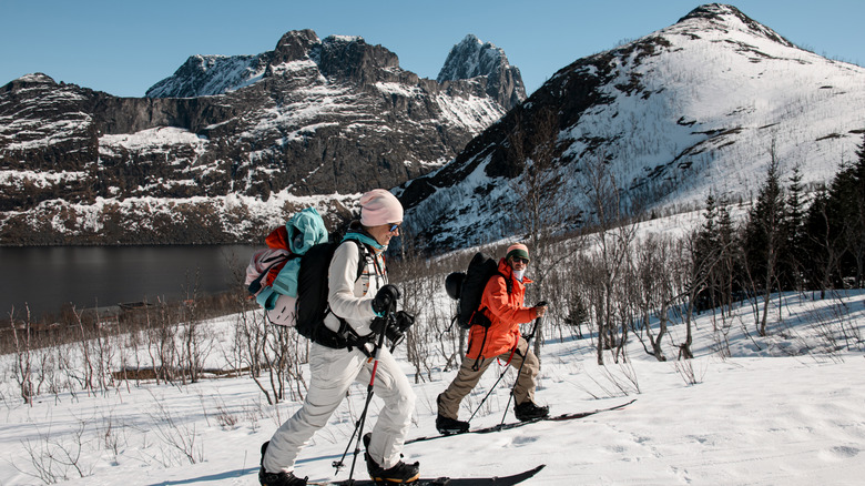 Side view of two skiers, a man and a smiling woman with a camera, ski up a snowy slope against the background of beautiful mountains and a body of water in the camera frame
